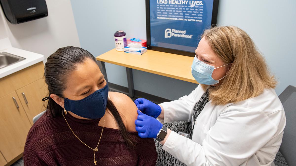 A Latina person gets a COVID-19 vaccination at Planned Parenthood North Central States. Both nurse and patient are wearing masks and smiling softly.