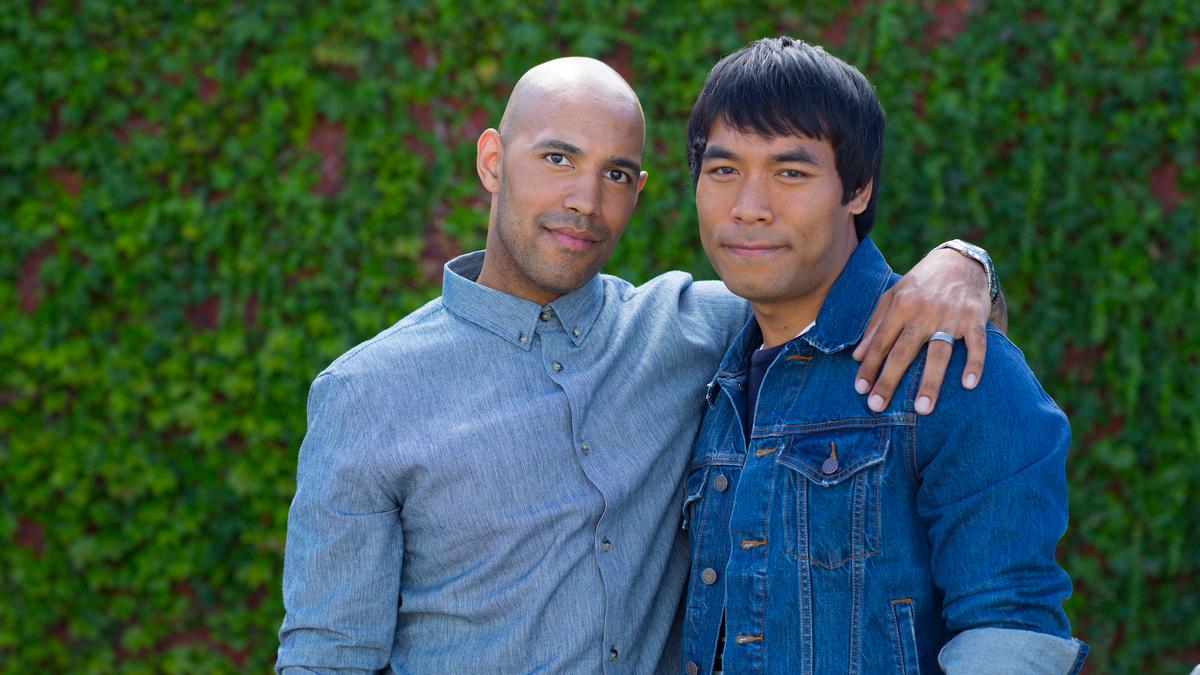 Two men in denim shirts in front of a greenery, one with his arm around the other, both looking at the camera.